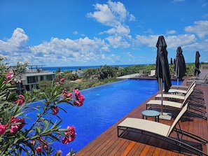 A swimming pool with lounge chairs and umbrellas, surrounded by tropical plants and a view of the ocean. - Private Whirlpool Terrace - Gated 3BR Playacar, Beach + Town (Playa del Carmen)