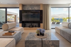 A modern living room with a flat-screen TV, comfortable seating, a chessboard, and a view of a body of water and mountains. - Villa on Brač, 80 m from the sea, with a gym, a heated pool, a boat and driver (Splitska)