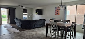 Ein Wohnzimmer mit einem dunklen Sofa, einem Esstisch mit Stühlen, einem Deckenventilator und einem an der Wand befestigten Fernseher.