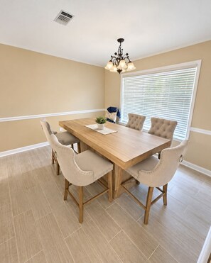 A dining room with a wooden table and six chairs, a chandelier, a window with blinds, and a plant on the table. - The Southern Standard, Sleeps 6 (Brandon)