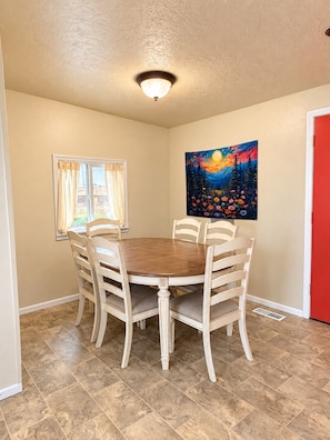 A dining area with a round table and six chairs, a window with curtains, a painting on the wall, and a red door. - Mountain and Meadows, family-friendly 3BR, near I-84, parks and dining. (Mountain Home)