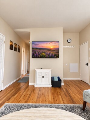 Een gang met een houten vloer, een wit kastje, een zwarte opbergkist, een klok aan de muur en een groot fotokader met een zonsondergang over een veld.