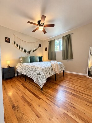 Un dormitorio con cama, piso de madera, ventilador de techo, mesita de noche con lámpara y ventana con cortinas.