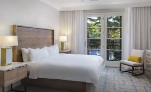 Ein Hotelzimmer mit einem großen Bett, zwei Nachttischen mit Lampen, einem Sessel mit Kissen und Blick auf Palmen durch eine Glastür.