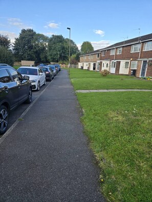 A row of parked cars along a paved path in a residential area with houses on one side and a grassy field on the other. - A Cozy 2 Bedroom Apartment Close to NEC & Airport (Birmingham)
