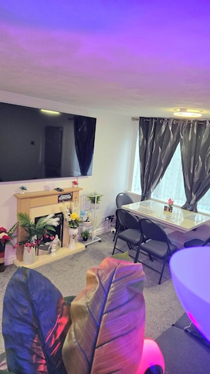 A room with a flat-screen TV mounted on the wall, a wooden cabinet with a sign, a dining table with chairs, and a window with curtains. - A Cozy 2 Bedroom Apartment Close to NEC & Airport (Birmingham)
