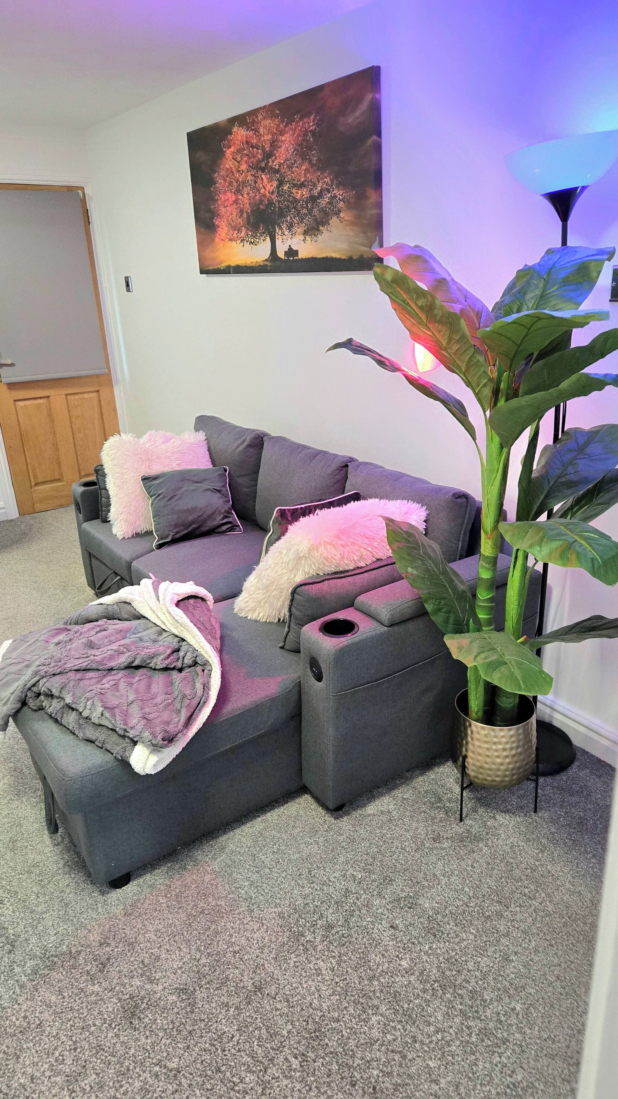 A grey sectional sofa with a pink throw blanket and pillows, a large potted plant, and a painting on the wall.