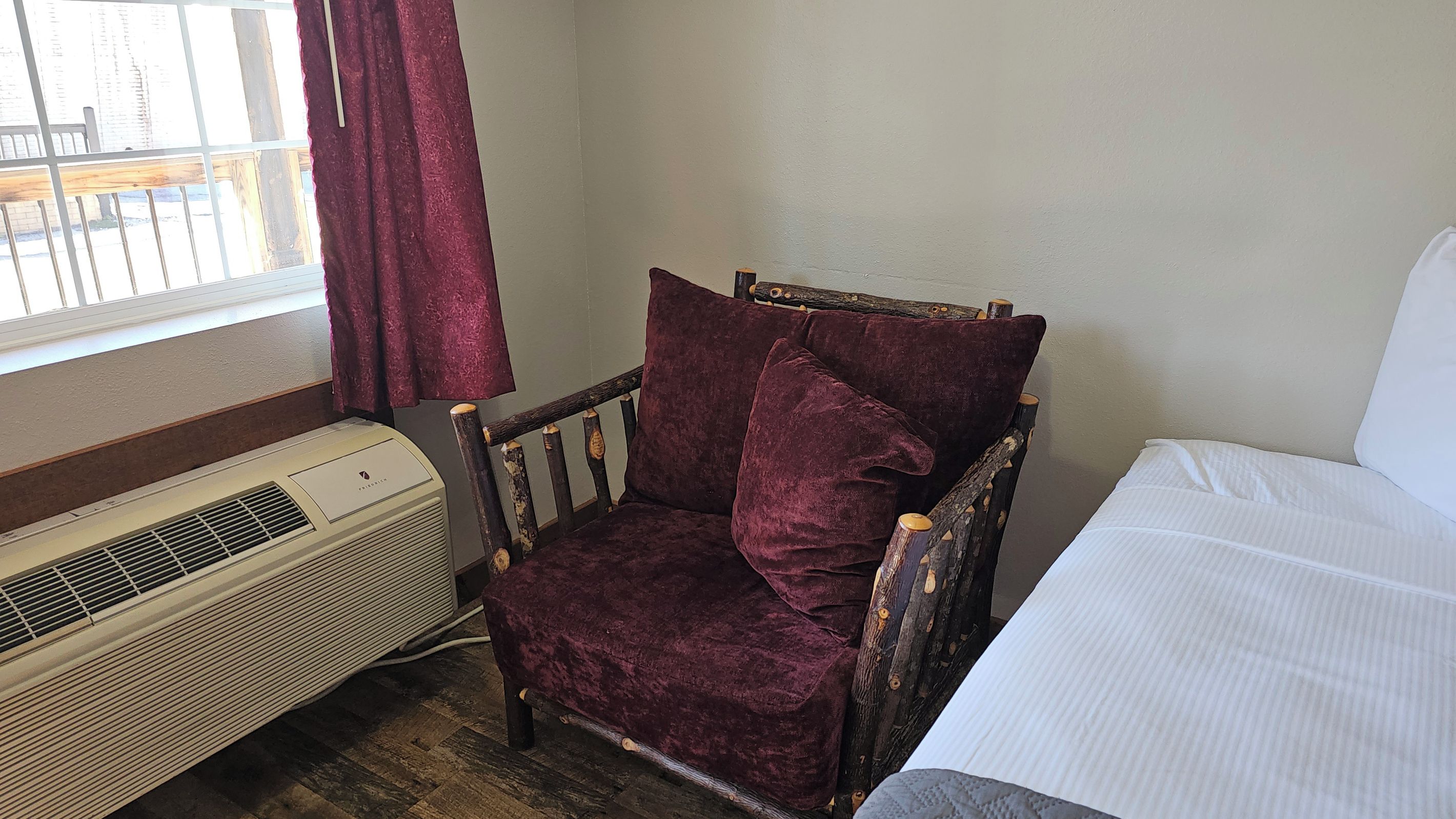 A room with a wooden chair with cushions, a white bed, and a window with curtains.