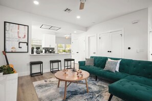 Una sala de estar moderna con un sofá verde, una mesa de centro redonda de madera y una cocina al fondo.