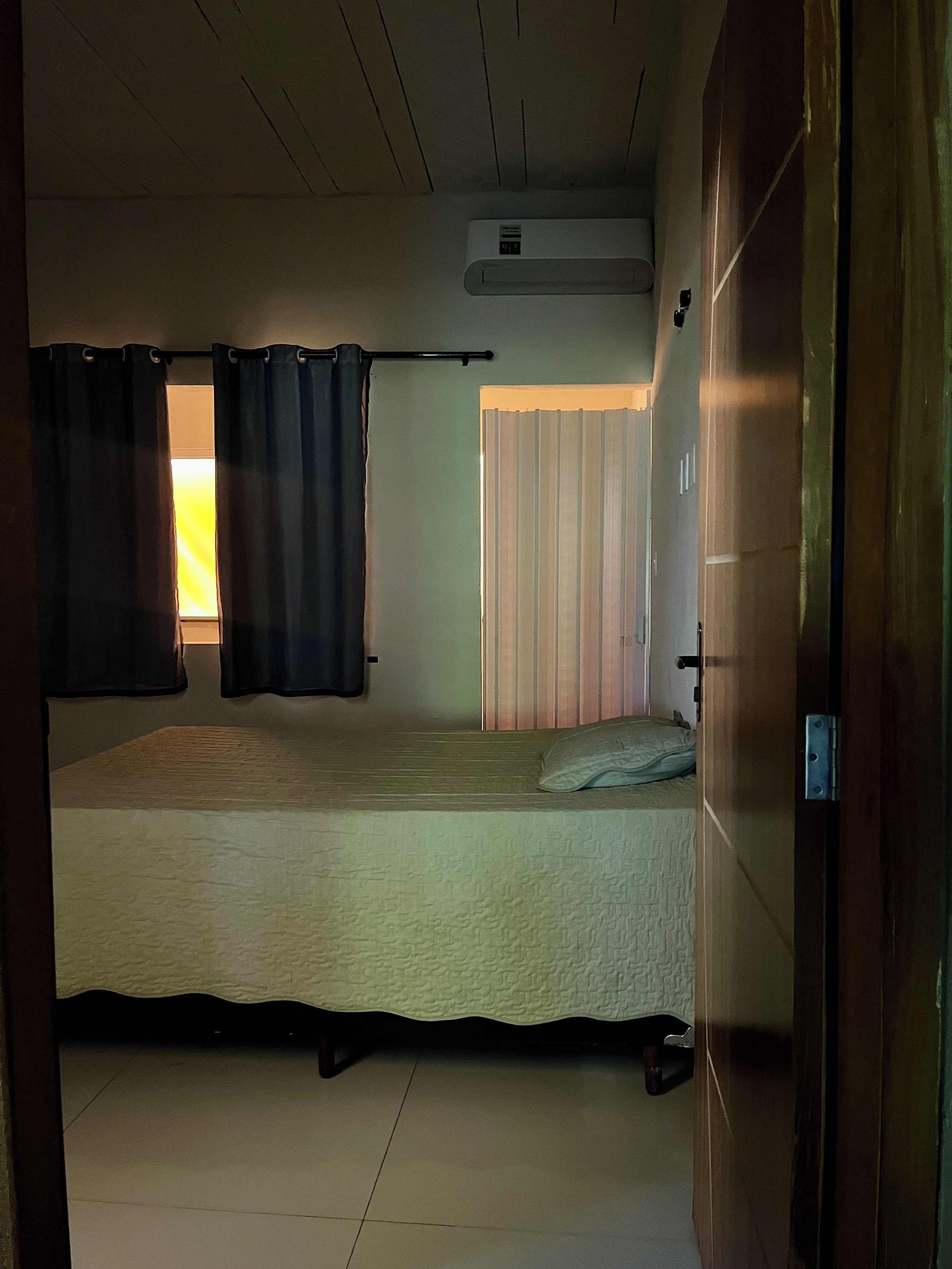 Een kamer met een bed, gordijn, raam met zonwering en een airconditioning.
