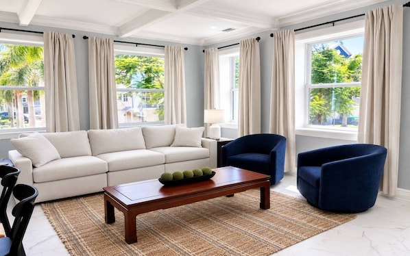 Ein modernes Wohnzimmer mit einer Couch, zwei Sesseln, einem Couchtisch und Blick auf Palmen durch große Fenster.