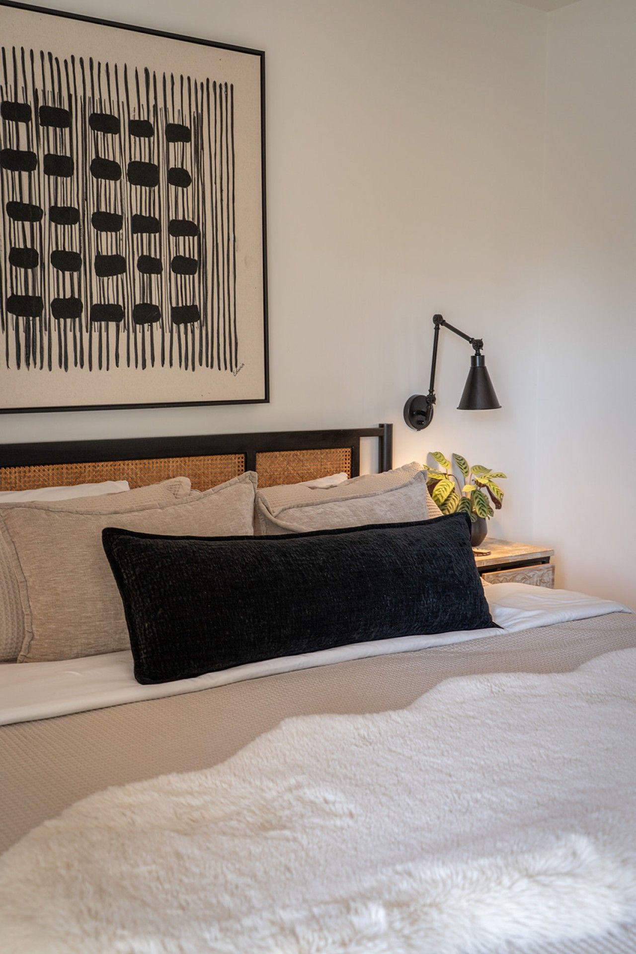 A neatly made bed with a black pillow, a headboard with a woven pattern, a wall-mounted abstract artwork, and a bedside table with a lamp and a potted plant.