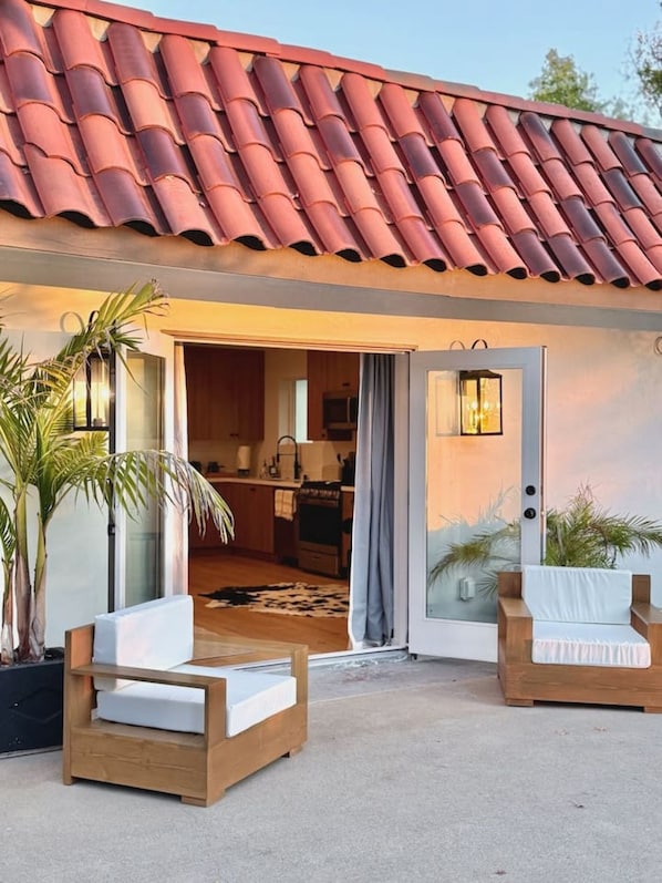 A modern house with a red-tiled roof, a glass door entrance, a kitchen visible through the doorway, and two wooden chairs with white cushions outside.