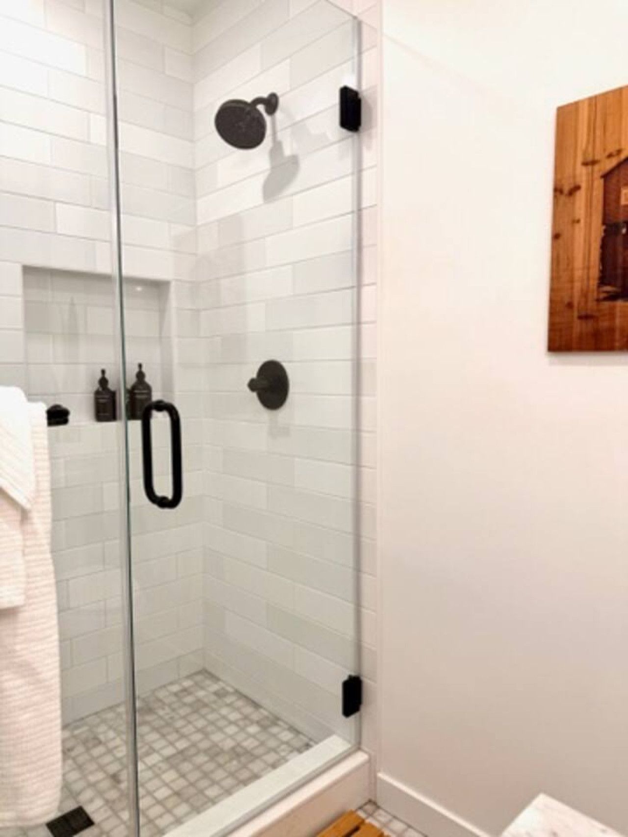 A glass-enclosed shower with white subway tiles, a black showerhead, and a wooden wall decoration.