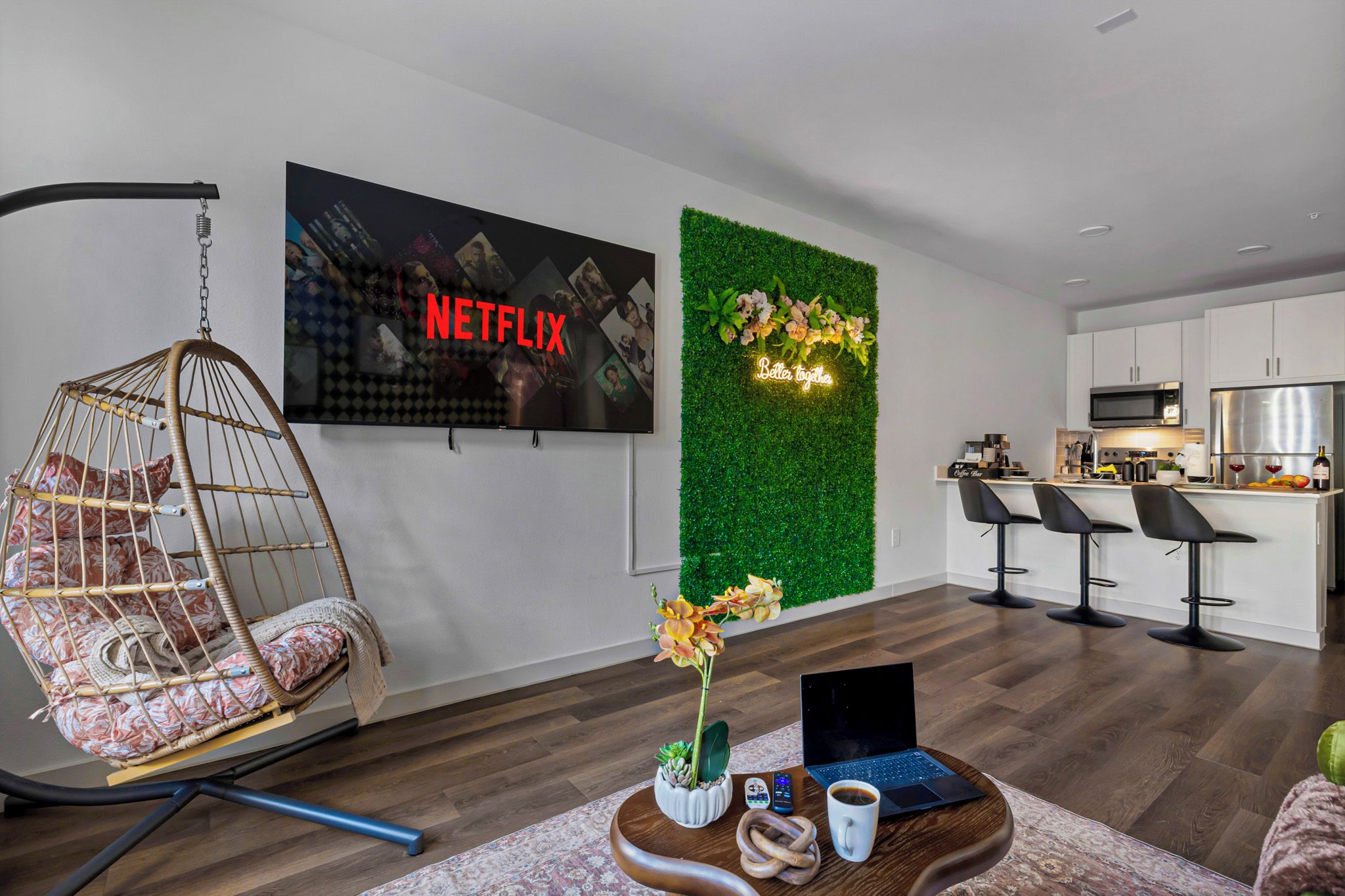 Un espace de vie moderne comprenant un fauteuil suspendu, un téléviseur fixé au mur affichant le logo « NETFLIX », un mur végétal agrémenté d’une composition florale et un coin cuisine avec des tabourets de bar.