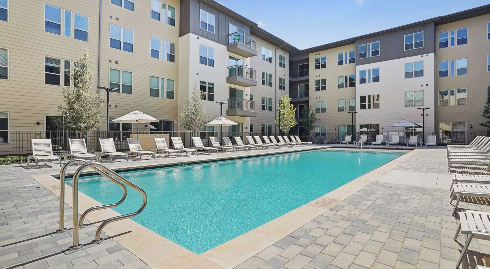A swimming pool with lounge chairs and umbrellas, surrounded by multi-story buildings with balconies.