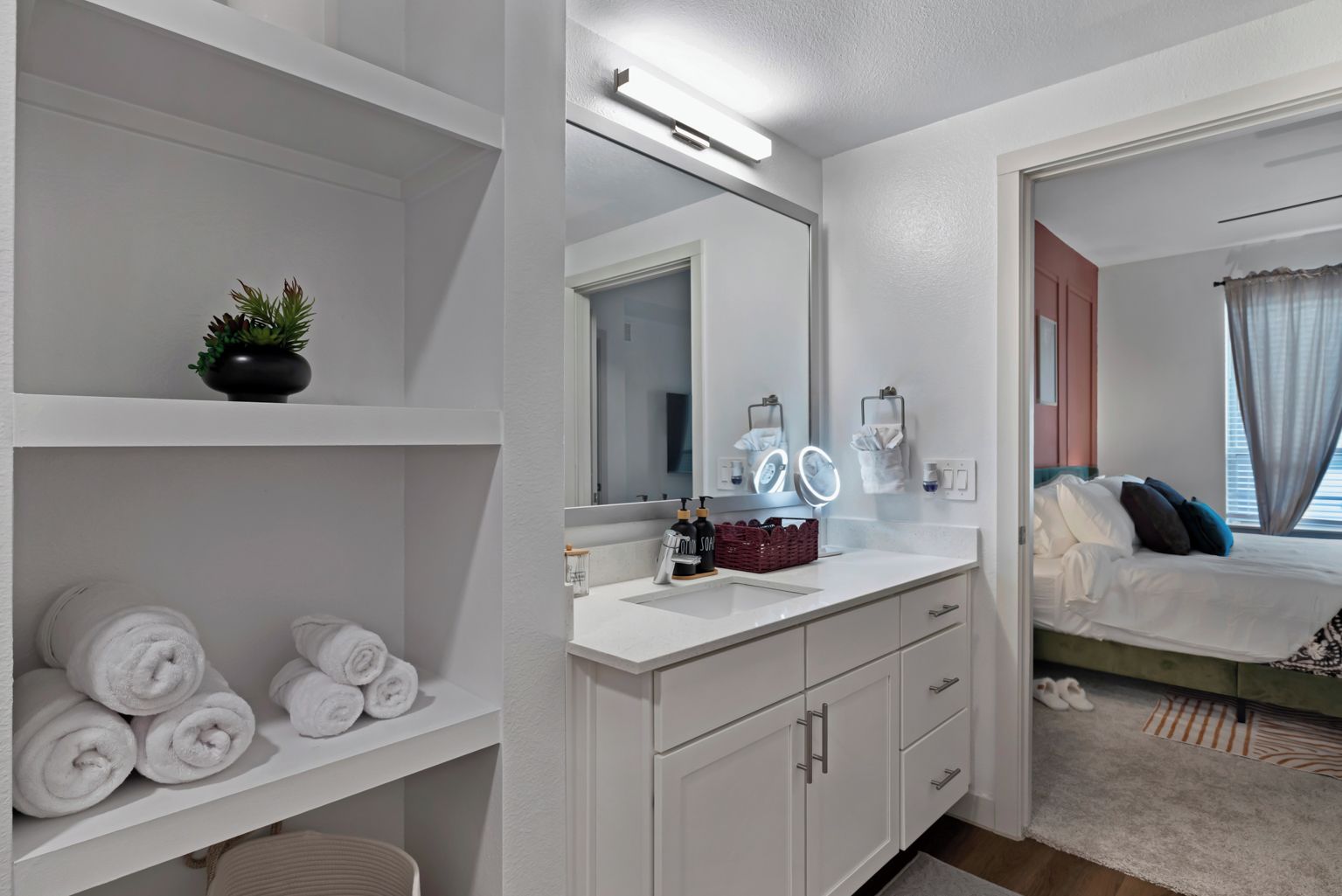 A modern bathroom with a white vanity, a large mirror, and a shelf with rolled towels.