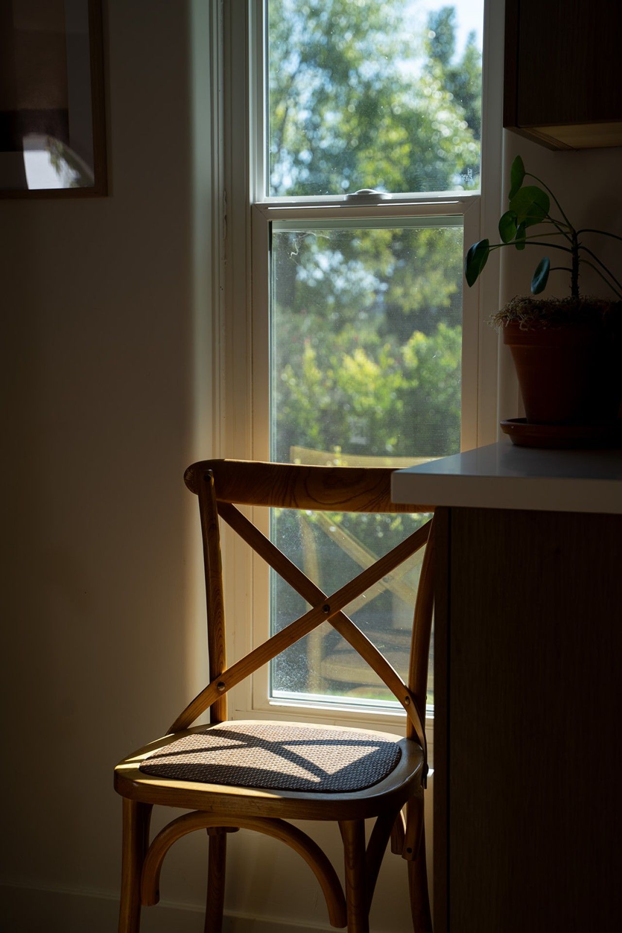 Una silla de madera con asiento de mimbre, ubicada junto a una ventana con vistas a árboles.