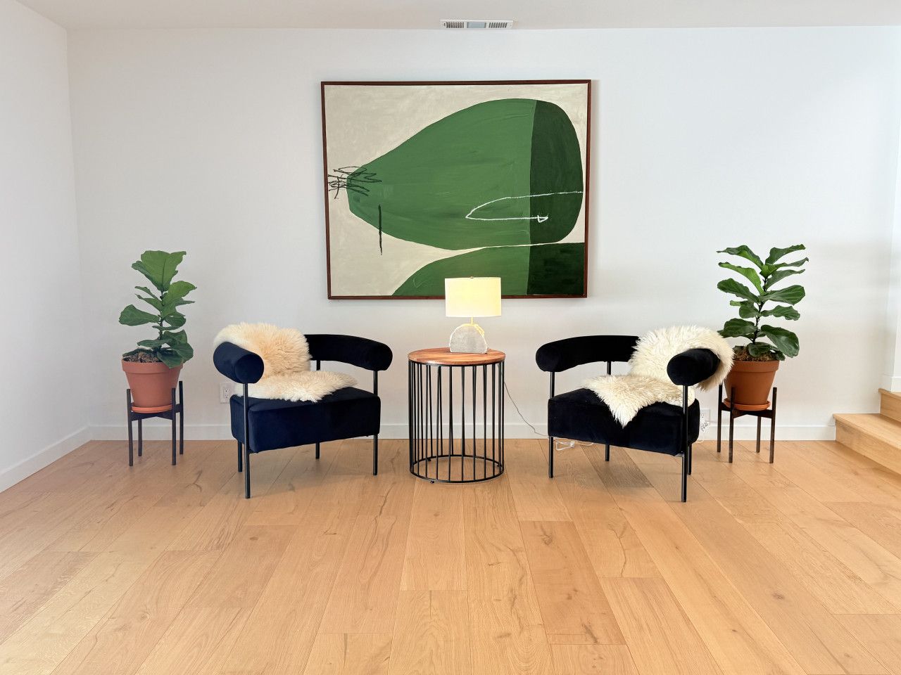 A room with two black chairs with fur throws, a round wooden table with a lamp, and two potted plants.