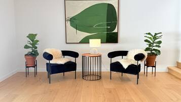 A room with two black chairs with fur throws, a round wooden table with a lamp, and two potted plants.