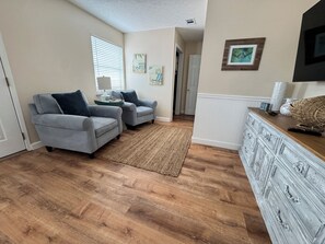 Ein Wohnzimmer mit zwei grauen Sesseln, Holzboden, einem gewebten Teppich und einer weißen Kommode mit Dekoration.