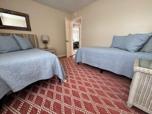 A room with two beds, a patterned red carpet, a small table with a lamp, and a wicker chair. - Walk to the Beach! Updated 3BR PCB Getaway for 6 (Panama City Beach)