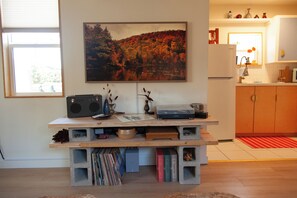 Quarto com escrivaninha de madeira, pintura de uma floresta e cozinha ao fundo.