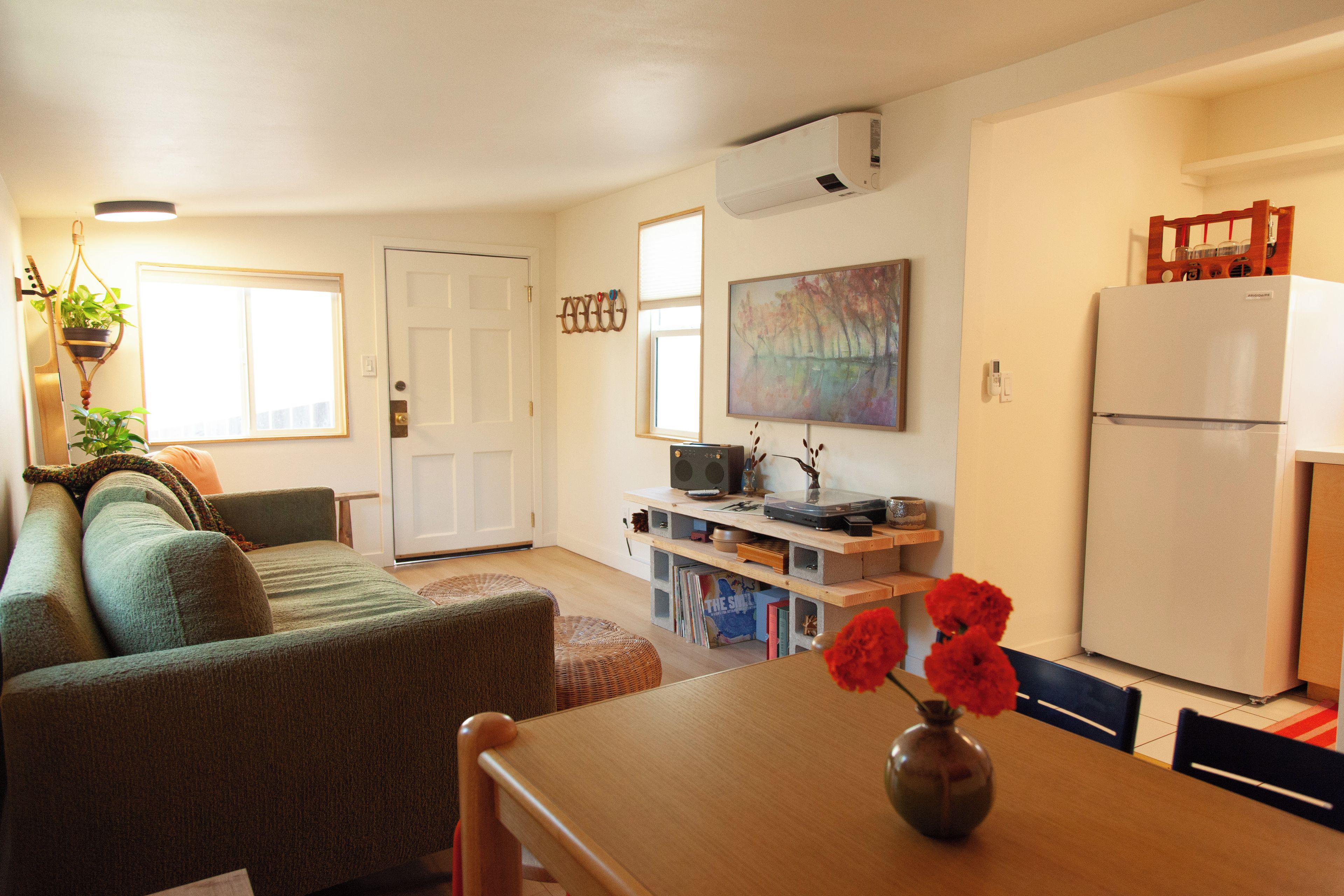 A compact living space with a sofa, a dining table with a vase, a kitchen area with a refrigerator, and a door leading to another room.