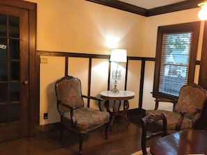 Quarto com duas poltronas estofadas, uma mesa redonda pequena com abajur e uma janela com persianas.