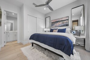 Ein Schlafzimmer mit einem Bett, einem Deckenventilator, einem Gemälde, einem Nachttisch und einem durch eine offene Tür einsehbaren Badezimmer.