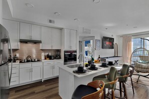 Une cuisine moderne avec des meubles blancs, un îlot central, des tabourets de bar et un fauteuil suspendu.