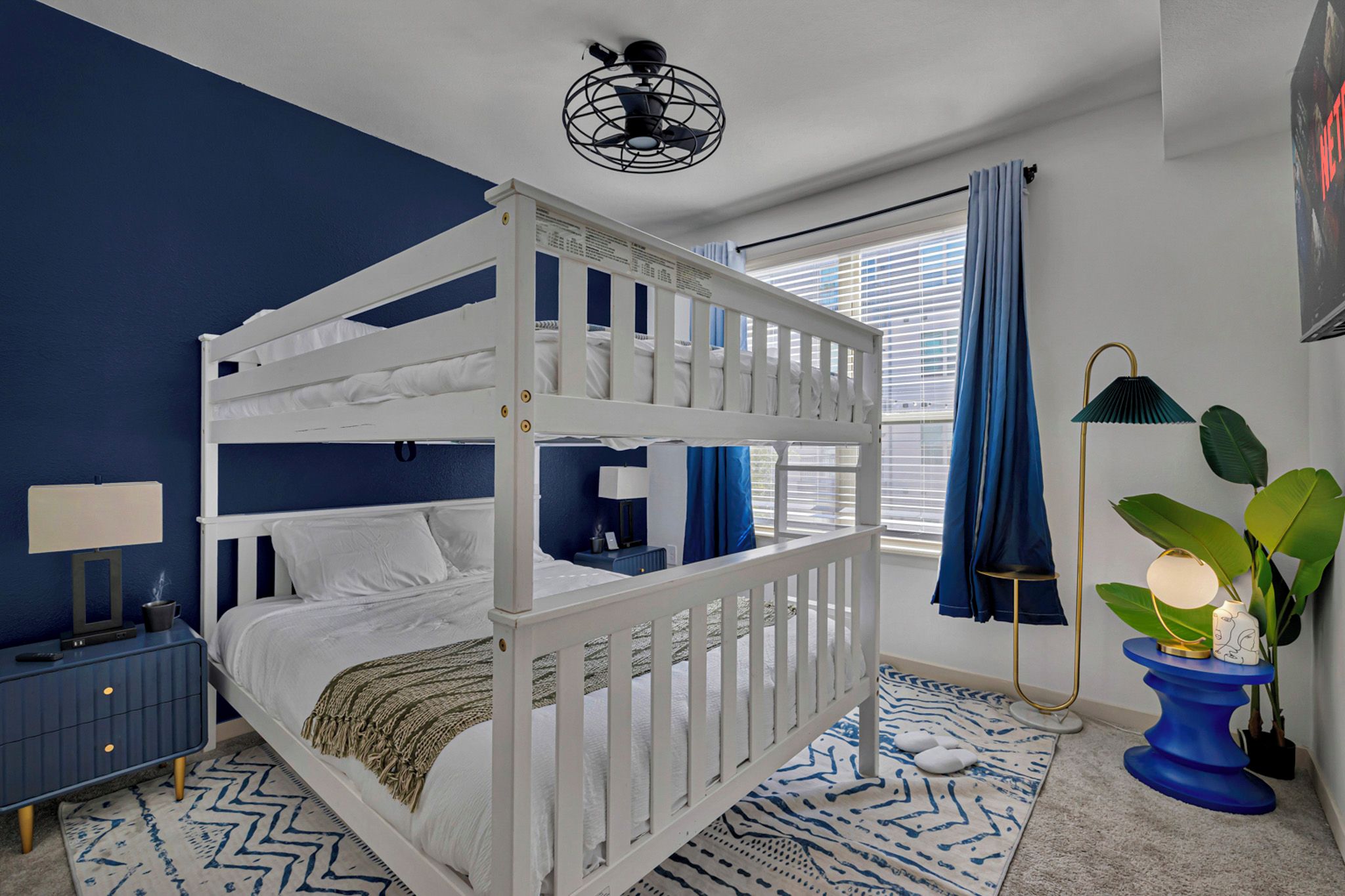 A bunk bed room with a blue and white color scheme, a bedside table with a lamp, a plant, and a window with curtains.