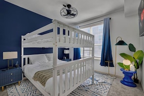 Une chambre avec un lit superposé aux couleurs bleu et blanc, une table de chevet avec une lampe, une plante et une fenêtre avec des rideaux.