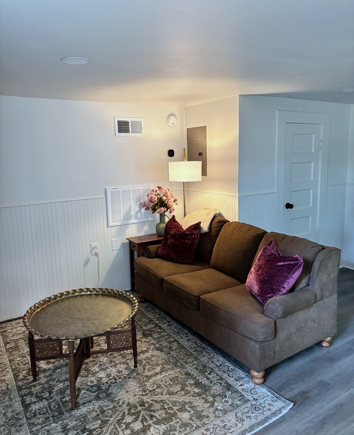 A room with a brown sofa, a round table, a lamp, and a door.