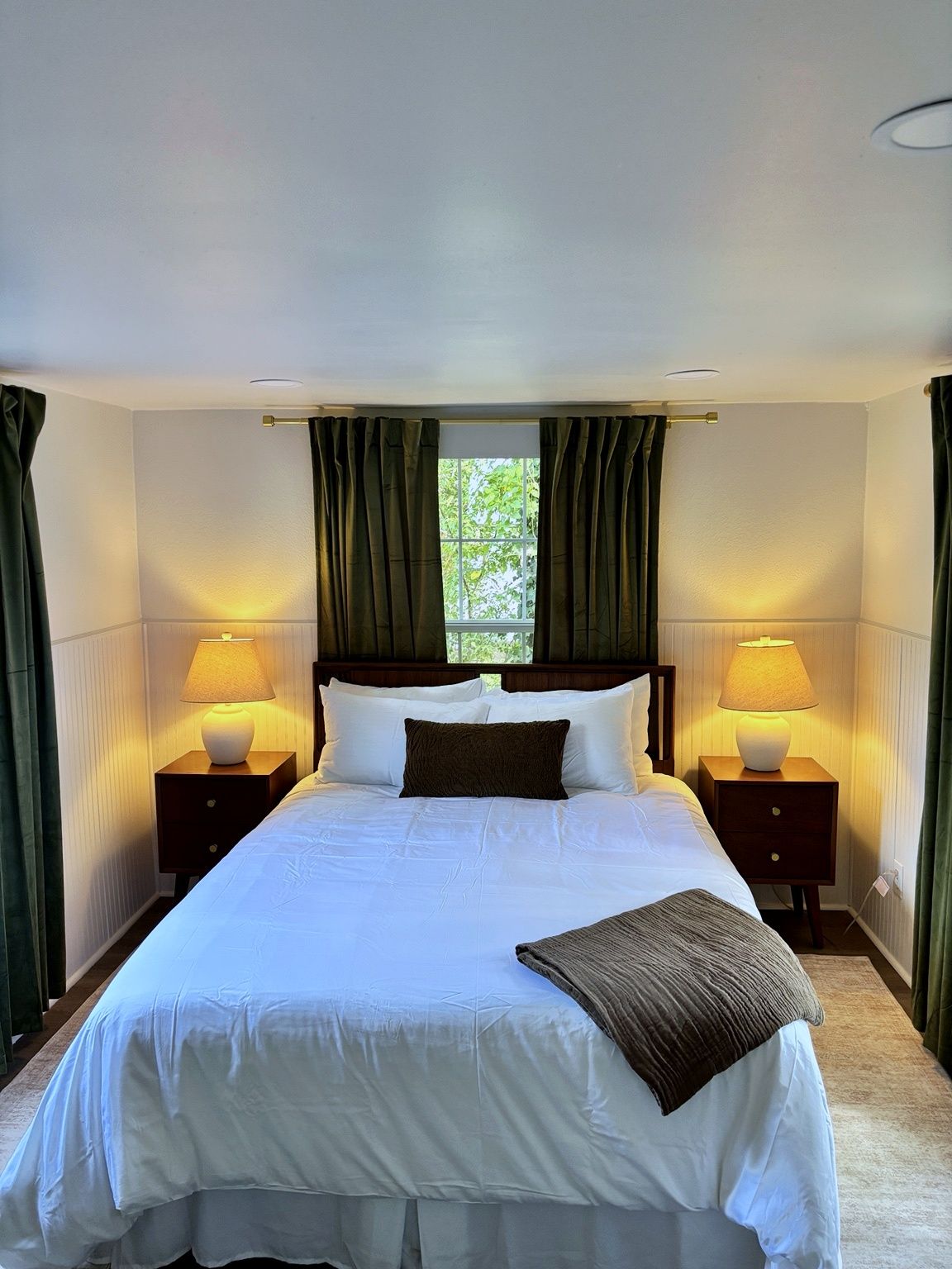 A hotel room with a large bed, two bedside tables with lamps, green curtains, and a window with a view of greenery.