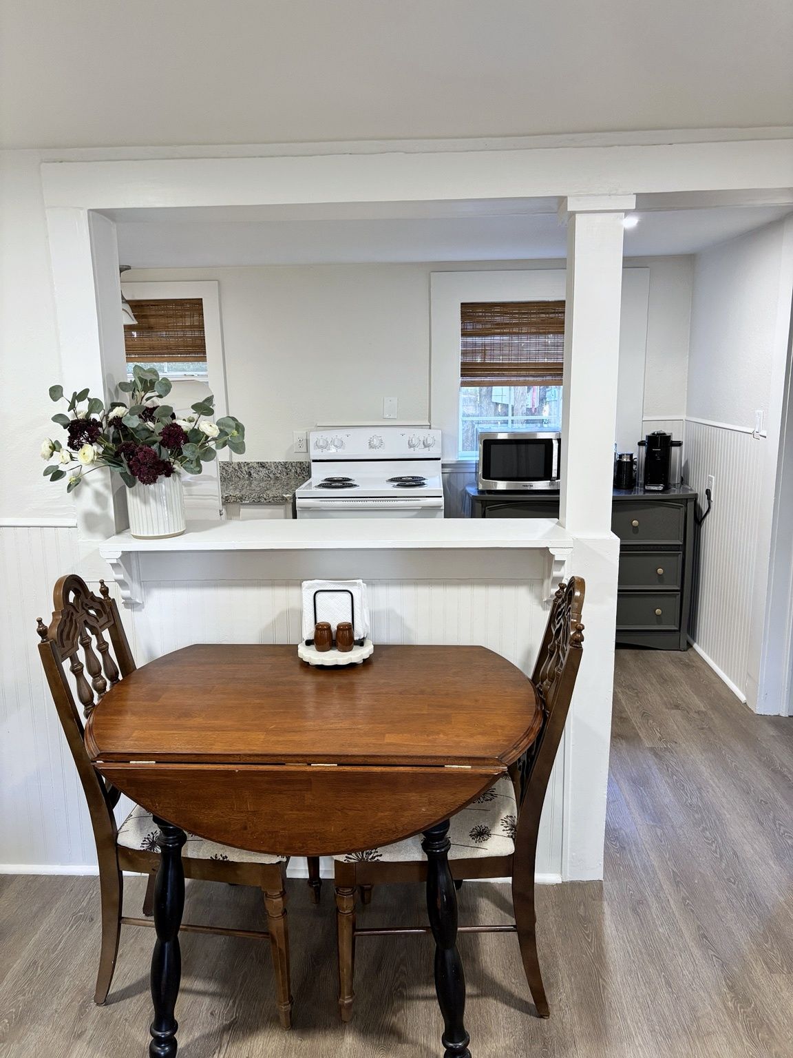 A dining area with a wooden table and chairs, a white kitchen in the background, and a window with blinds.