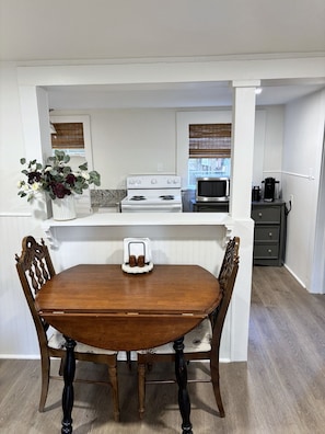 A dining area with a wooden table and chairs, a white kitchen in the background, and a window with blinds.