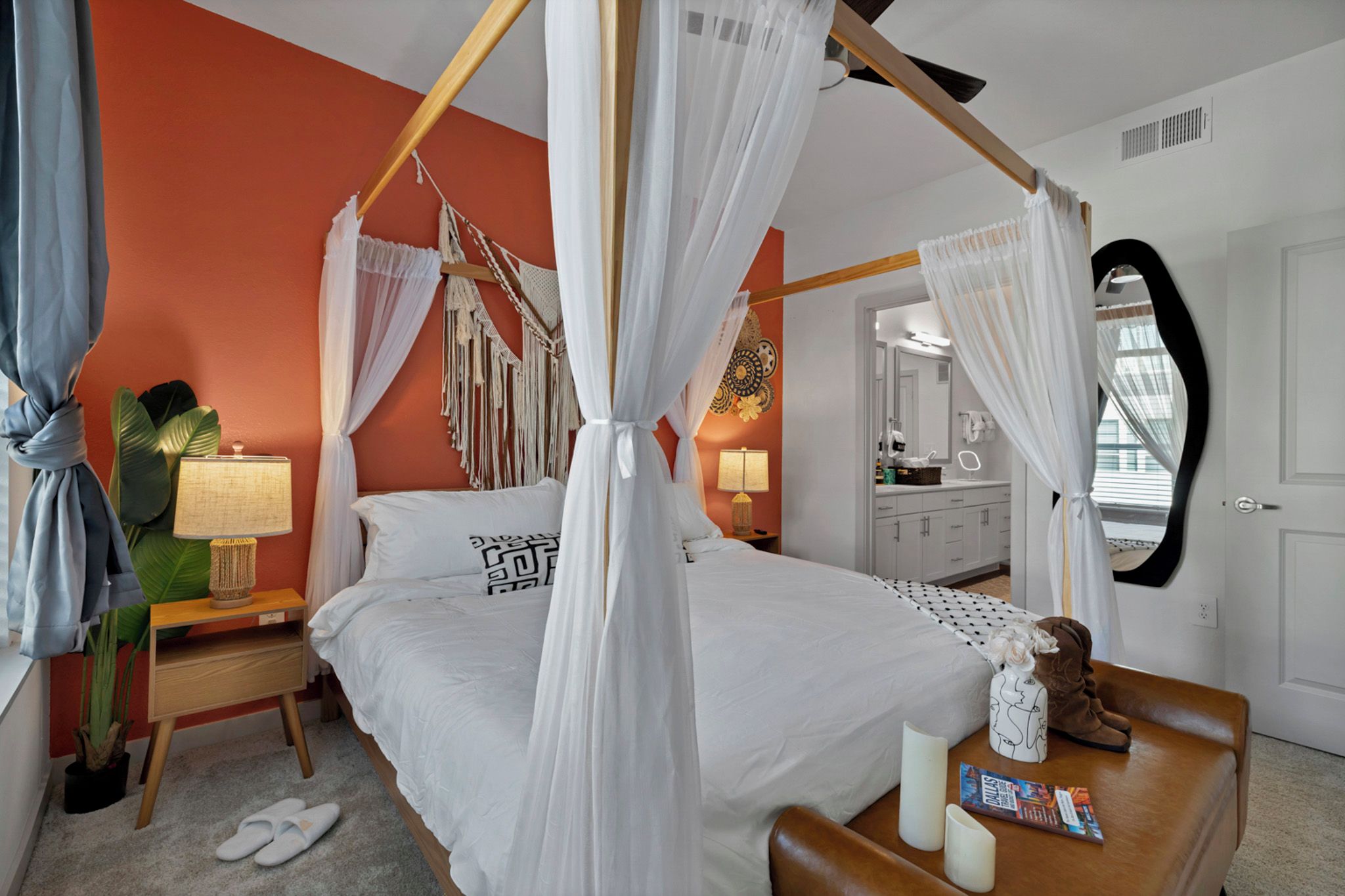 A four-poster canopy bed with a white bedspread and a geometric pillow design, a bedside table with a lamp, and a nightstand with a plant.