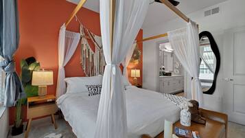 A four-poster canopy bed with a white bedspread and a geometric pillow design, a bedside table with a lamp, and a nightstand with a plant.