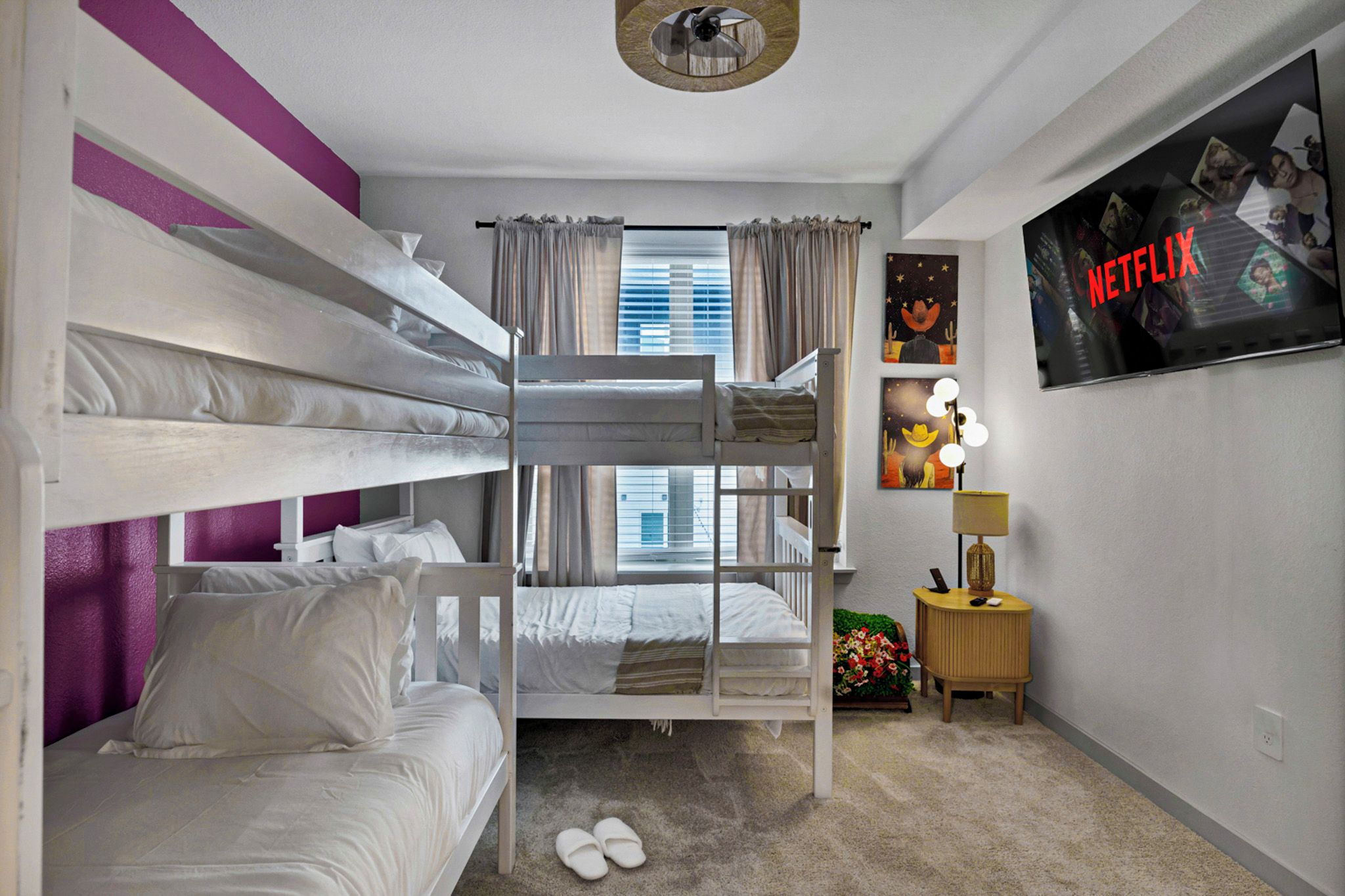 Une chambre avec un lit superposé, un téléviseur fixé au mur, une petite table avec une lampe et une fenêtre avec des rideaux.