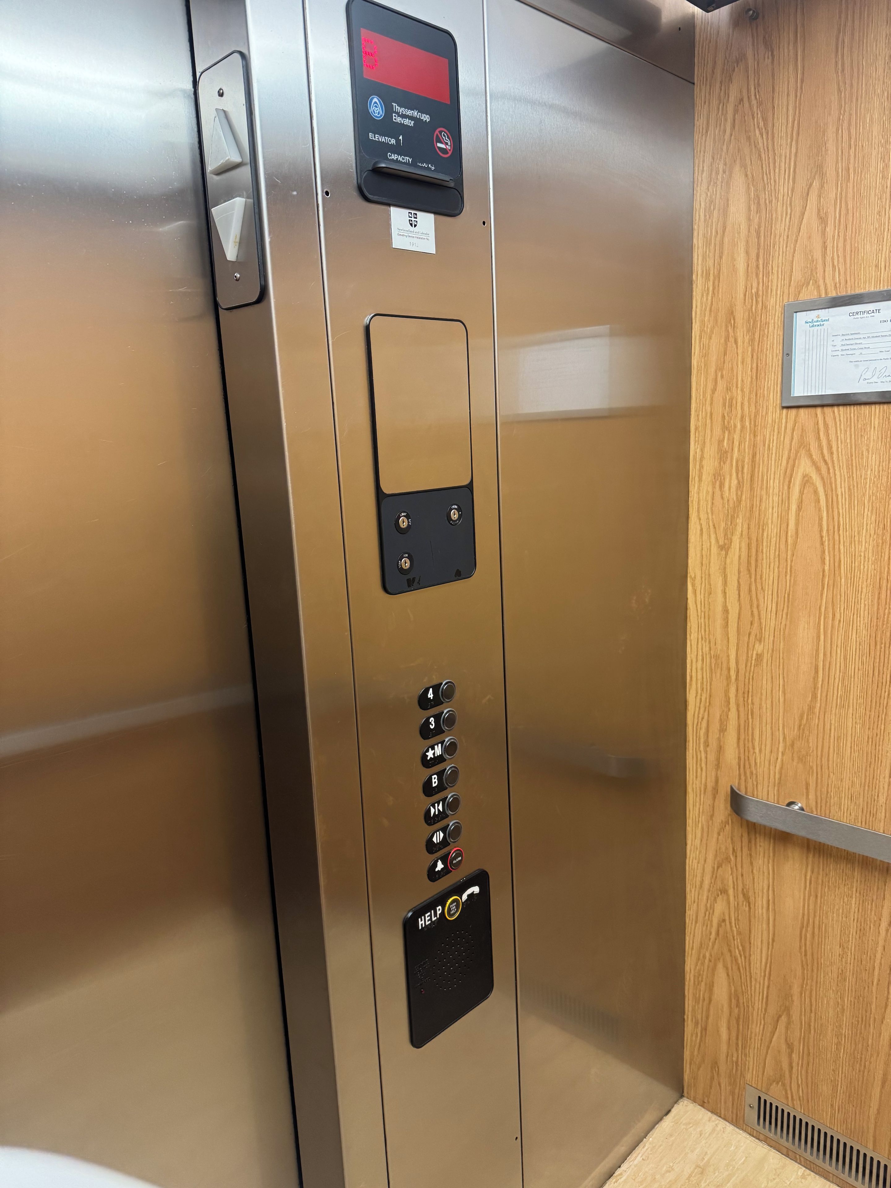 Interior de un ascensor con panel de control, certificado y puerta de madera.