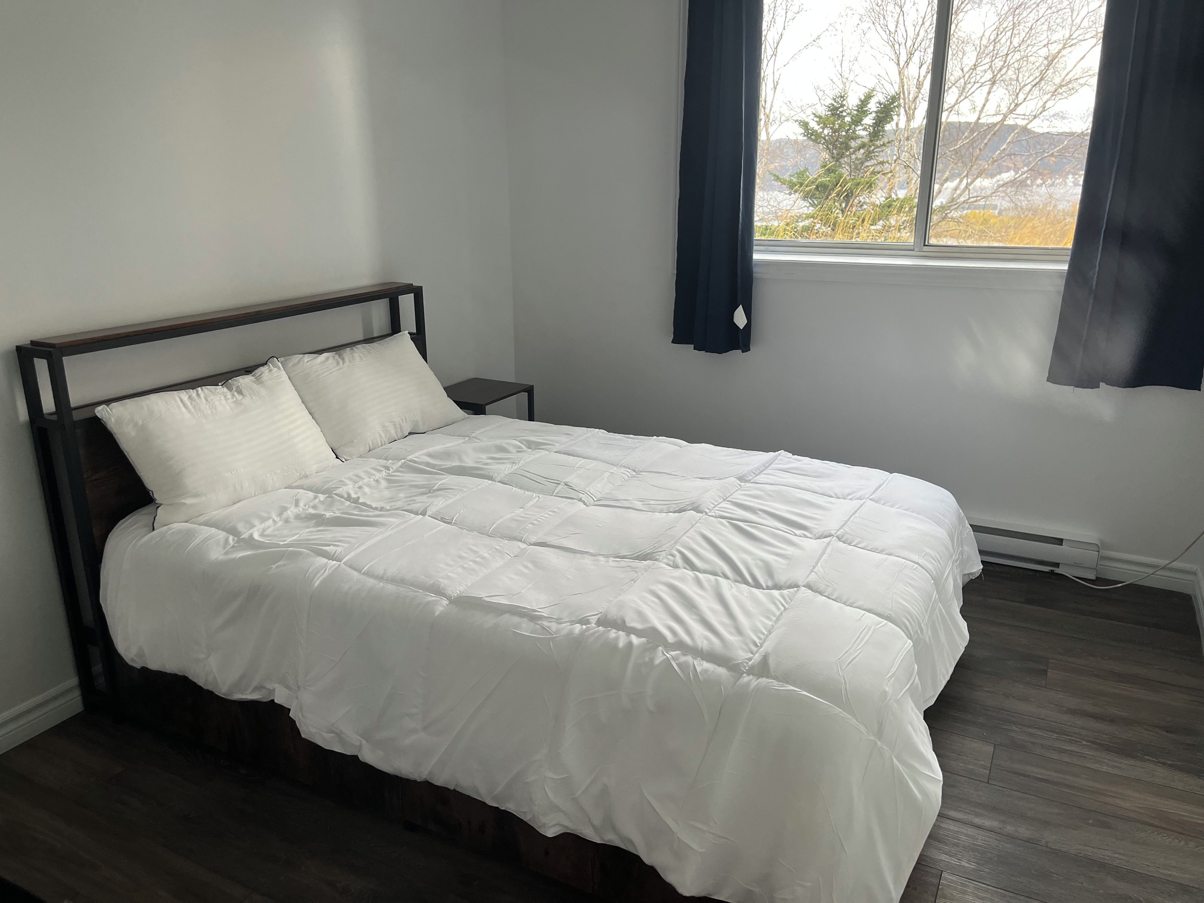 A bed with a wooden frame and white bedding in a room with a window and a small bedside table.