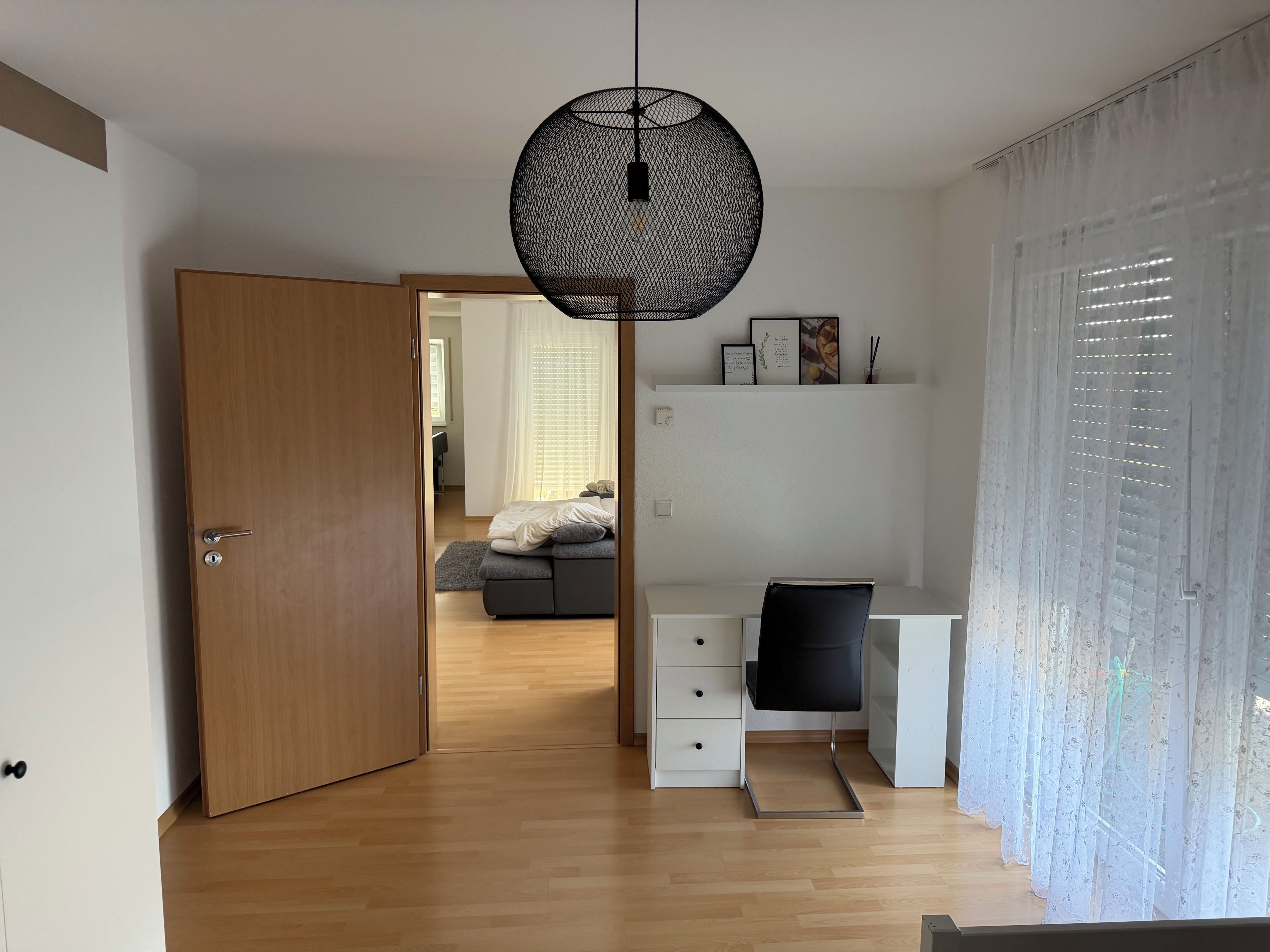 A modern bedroom with a wooden door, a desk with a chair, a bed with a gray blanket, and a hanging light fixture.