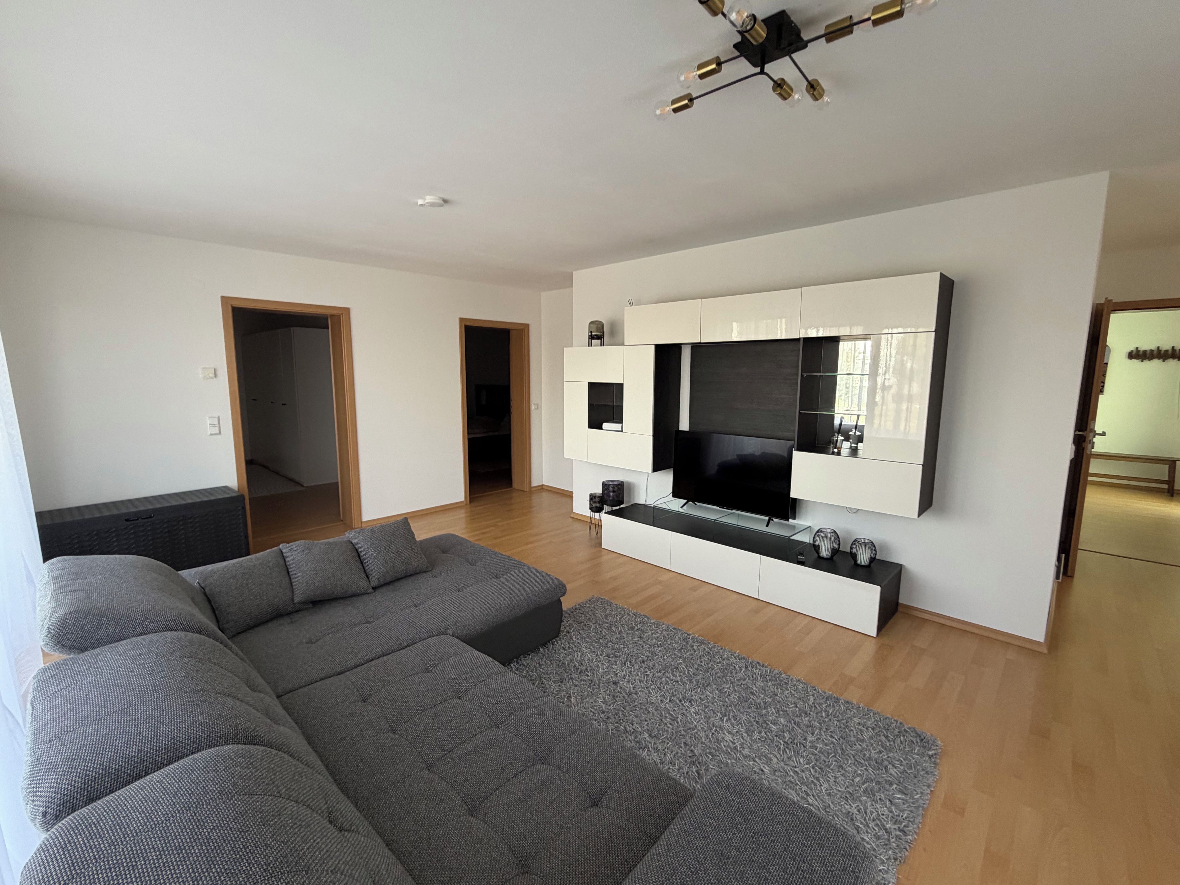 Ein modernes Wohnzimmer mit einer großen Eckcouch, einem Flachbildfernseher an einer weißen Wand und Holzboden.