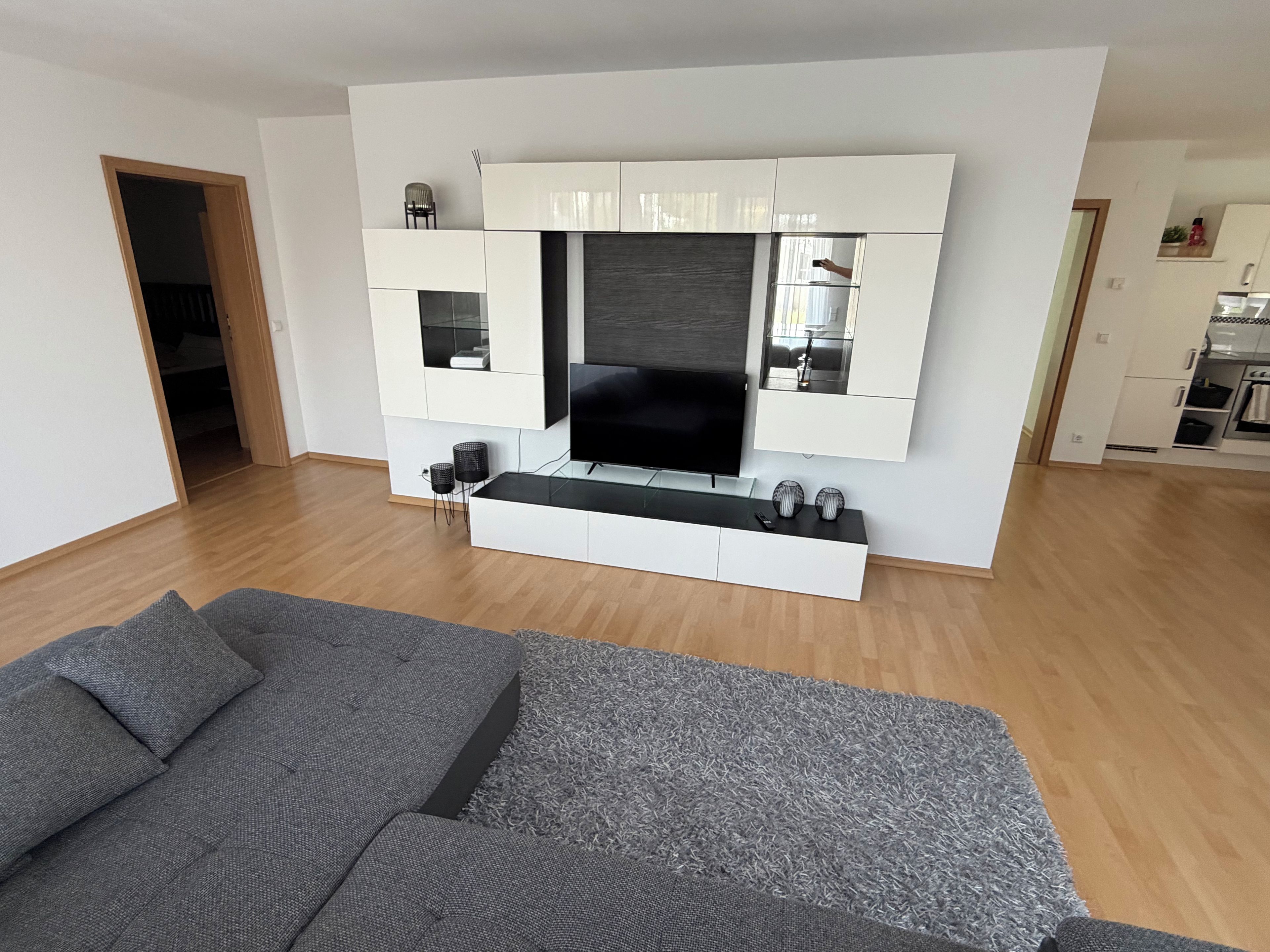 A modern living room with a flat-screen TV, built-in storage cabinets, a sofa, and a small kitchen area in the background.