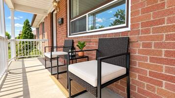 A covered outdoor seating area with two chairs and a table, a potted plant, and a window with a view of trees.