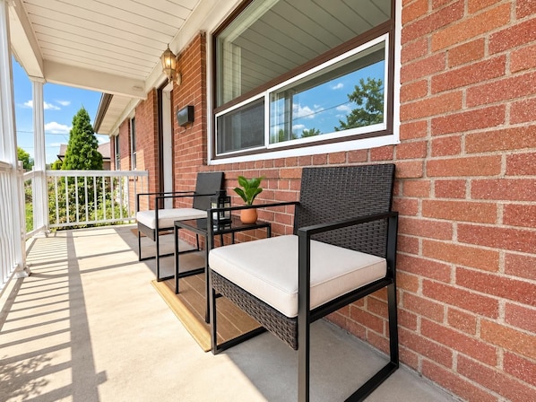 A covered outdoor seating area with two chairs and a table, a potted plant, and a window with a view of trees.