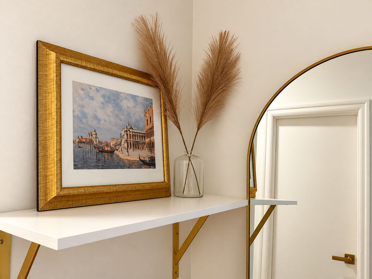 A framed picture of a cityscape on a white shelf, a vase with dried pampas grass, and a white door with a gold frame.