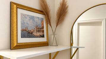 A framed picture of a cityscape on a white shelf, a vase with dried pampas grass, and a white door with a gold frame.