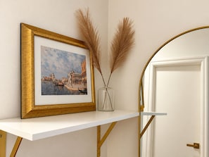 A framed picture of a cityscape on a white shelf, a vase with dried pampas grass, and a white door with a gold frame.